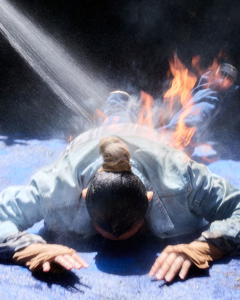Person wearing a denim jacket on fire lying facedown on a blue mat while being sprayed with water