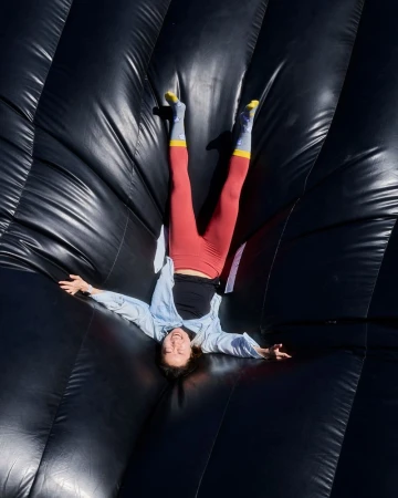 Smiling woman in red leggings and light blue shirt lying upside down on an inflated black stunt bag