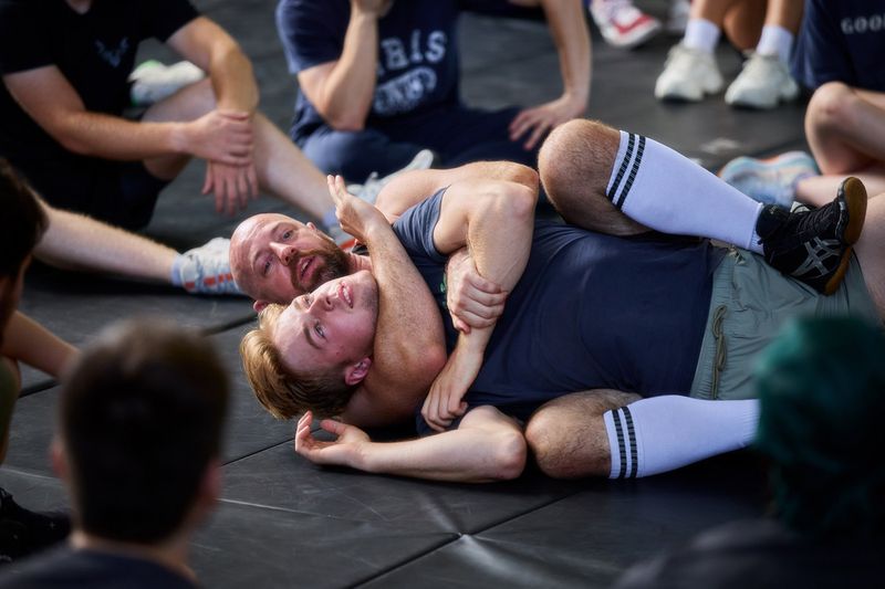 Two men grappling on a dark padded mat surrounded by seated observers during a stunt training session.