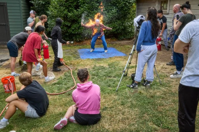 Person engulfed in flames standing on a blue mat surrounded by onlookers and crew members in an outdoor yard.