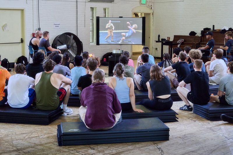 Group of students sitting on gym mats watching a video screen in a bright hall with exposed brick walls