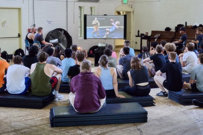 Group of students sitting on gym mats watching a video screen in a bright hall with exposed brick walls
