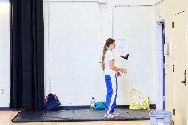Woman in white t-shirt and blue tracksuit trousers standing in a gymnasium holding a prop gun.