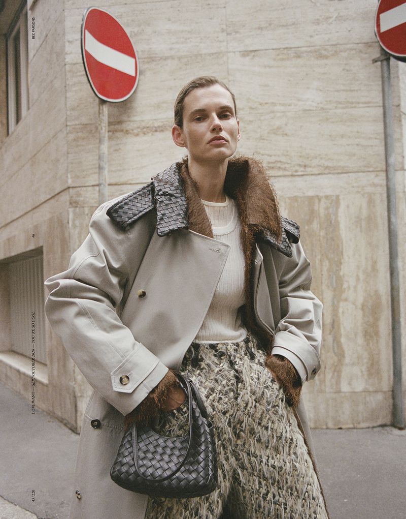 Model Giedre Dukauskaite wearing a beige trench coat and holding a woven black handbag on a city street.