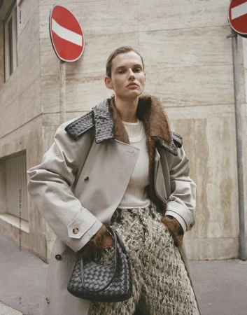 Model Giedre Dukauskaite wearing a beige trench coat and holding a woven black handbag on a city street.