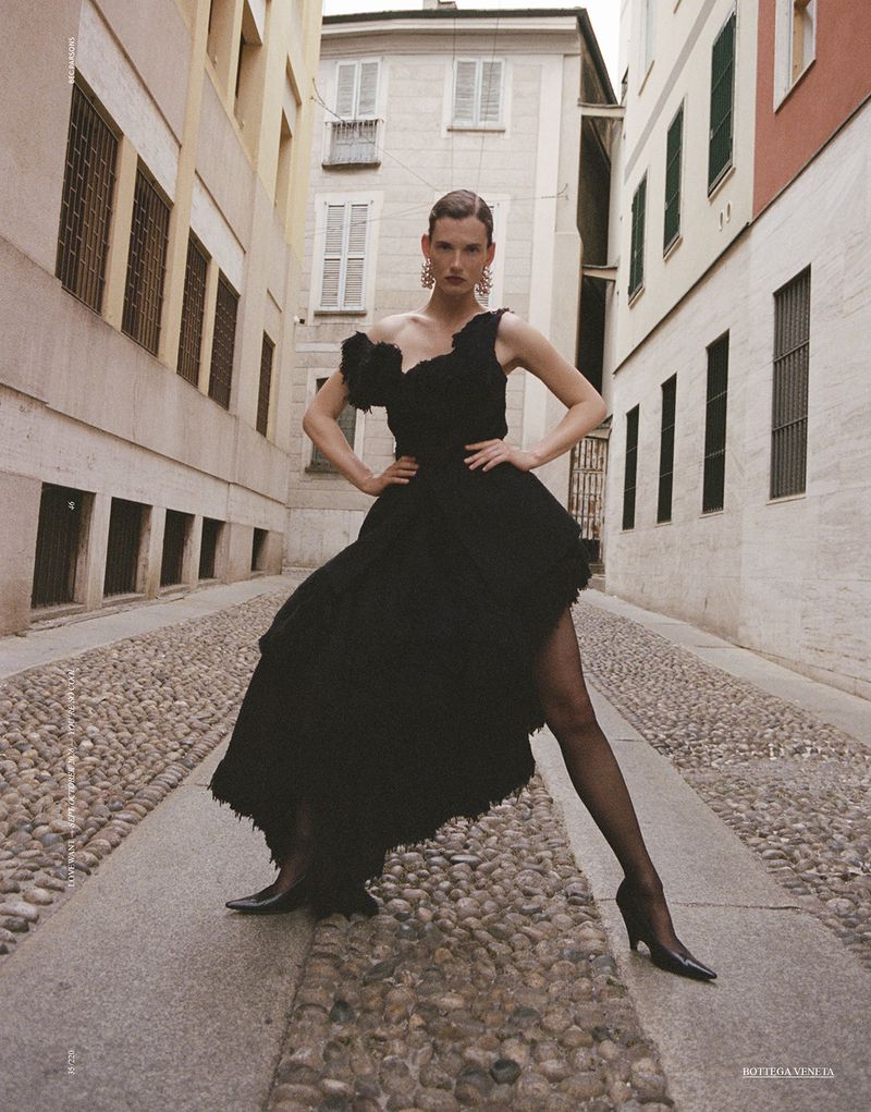 Model Giedre Dukauskaite in a black ruffled dress standing on a narrow cobblestone street lined with tall buildings.