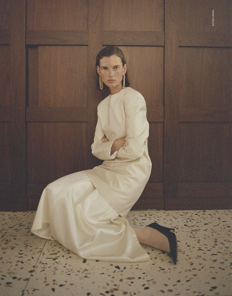 Model Giedre Dukauskaite seated on a terrazzo floor wearing a cream dress against wood paneling for Love Want Magazine.