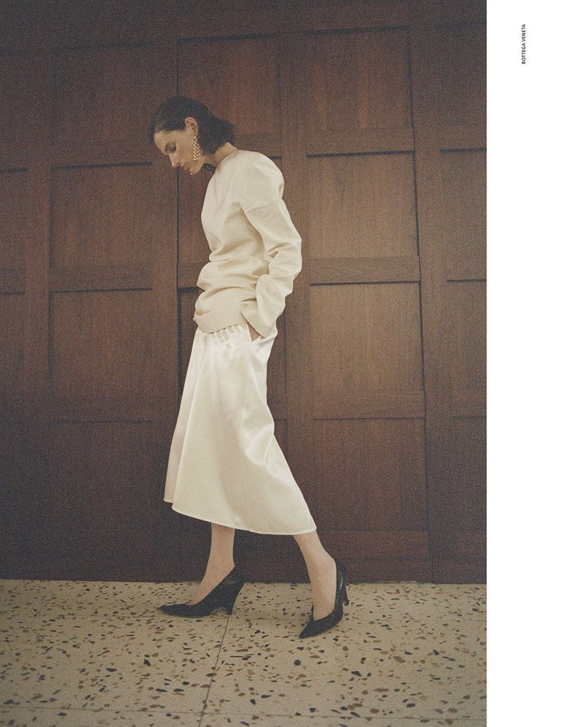 Model Giedre Dukauskaite in cream top and white skirt standing against dark wood paneling for Love Want Magazine.