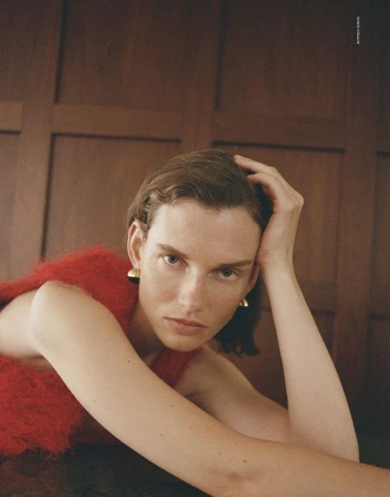 Model Giedre Dukauskaite wearing a red textured top and gold earrings, posing against wood paneling for Love Want Magazine.