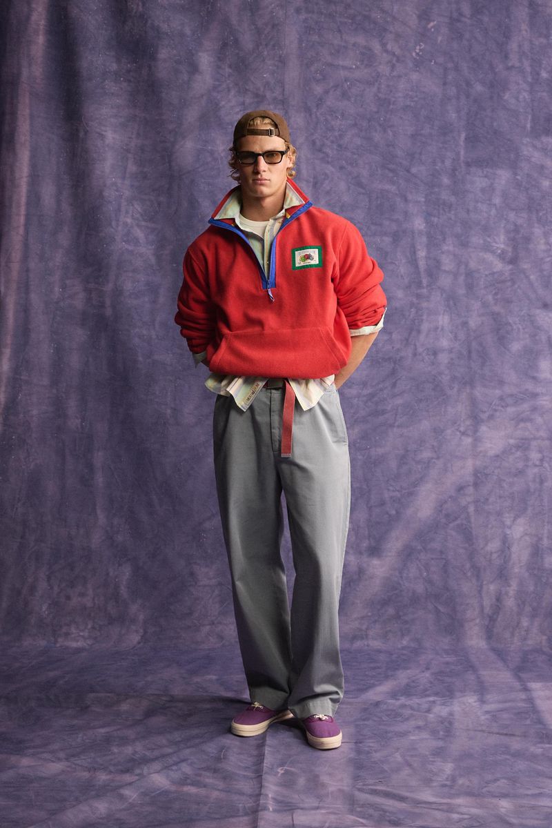 Male model in a red fleece quarter-zip pullover and grey trousers standing against a textured purple backdrop.