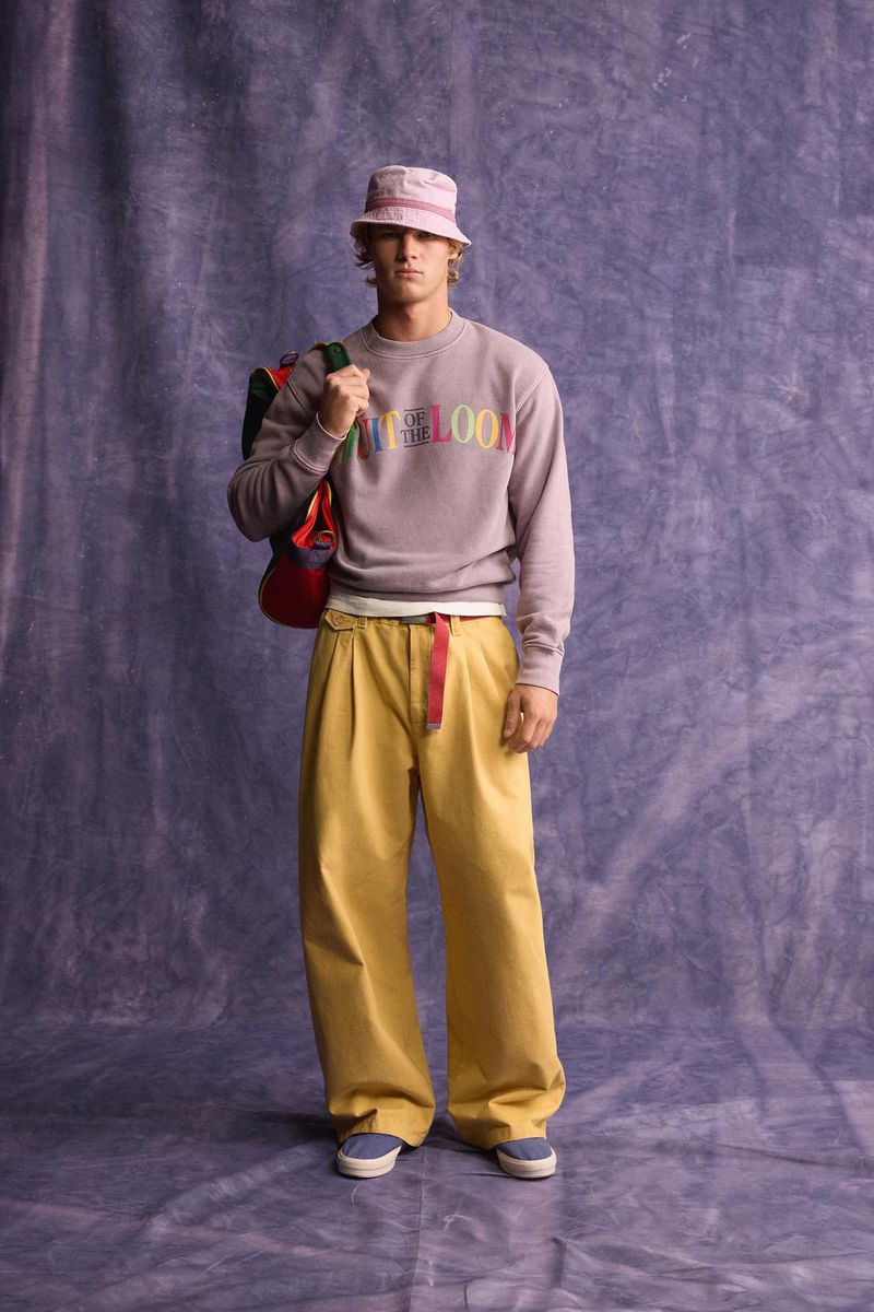 Male model wearing a lavender sweatshirt, yellow trousers, and pink bucket hat with a backpack against a textured backdrop