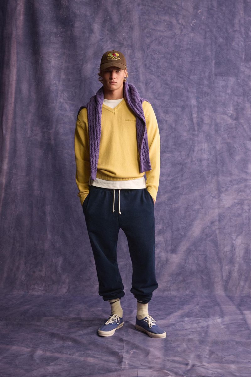 Young male model wearing yellow sweater, dark trousers, and a baseball cap against a textured purple backdrop.