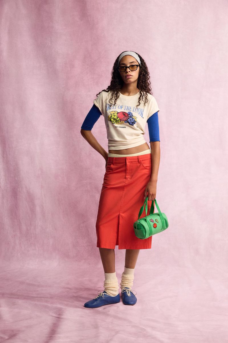 Model standing against a pink textured backdrop wearing a graphic t-shirt, blue sleeves, and a red skirt while holding a green handbag.