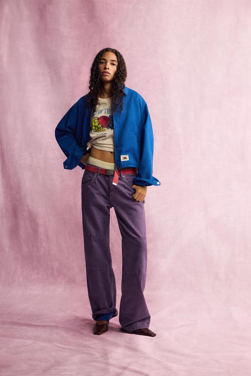 Model standing against a pink textured backdrop wearing a blue overshirt, graphic t-shirt, and purple trousers.