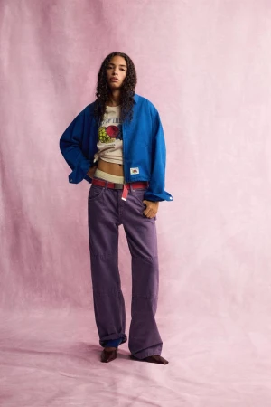 Model standing against a pink textured backdrop wearing a blue overshirt, graphic t-shirt, and purple trousers.