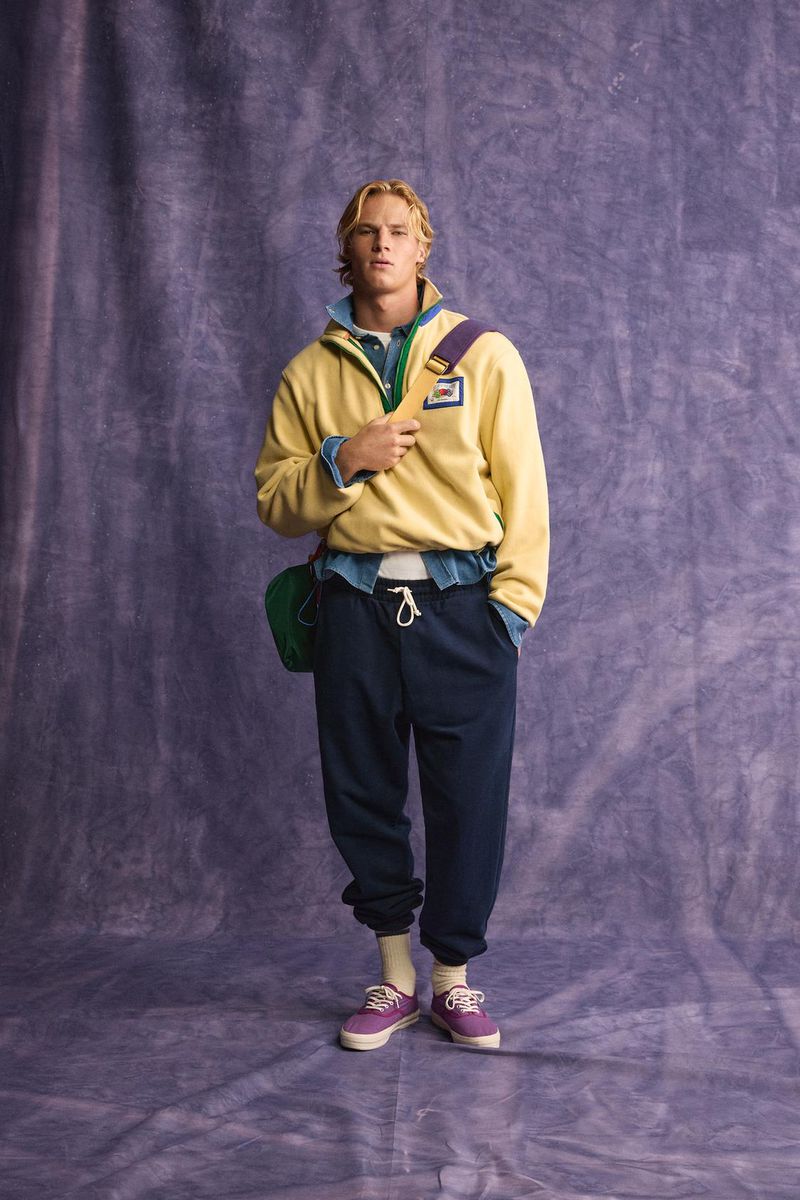 Model wearing a yellow fleece half-zip sweatshirt and dark navy sweatpants standing against a textured purple backdrop.