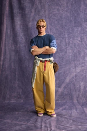 Model standing with arms crossed wearing a blue t-shirt and yellow trousers against a mottled purple background