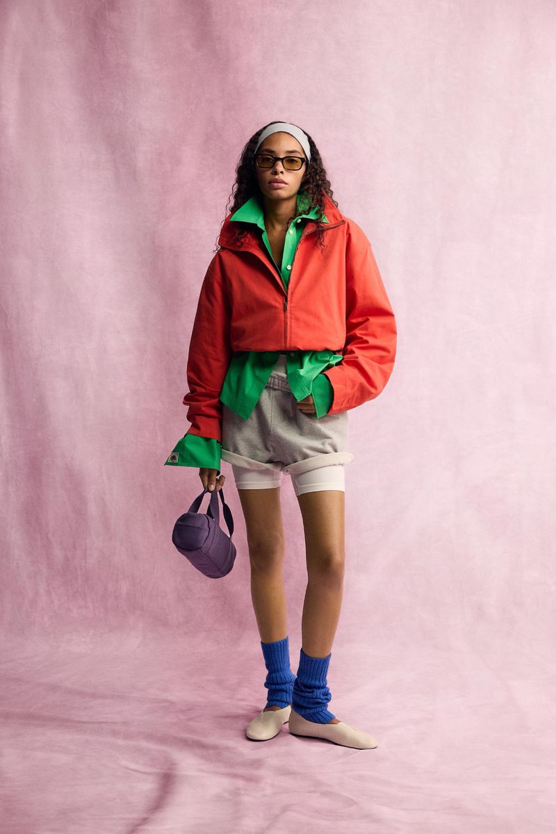 Model in red jacket and green shirt wearing blue leg warmers holding a purple handbag against a pink textured background.