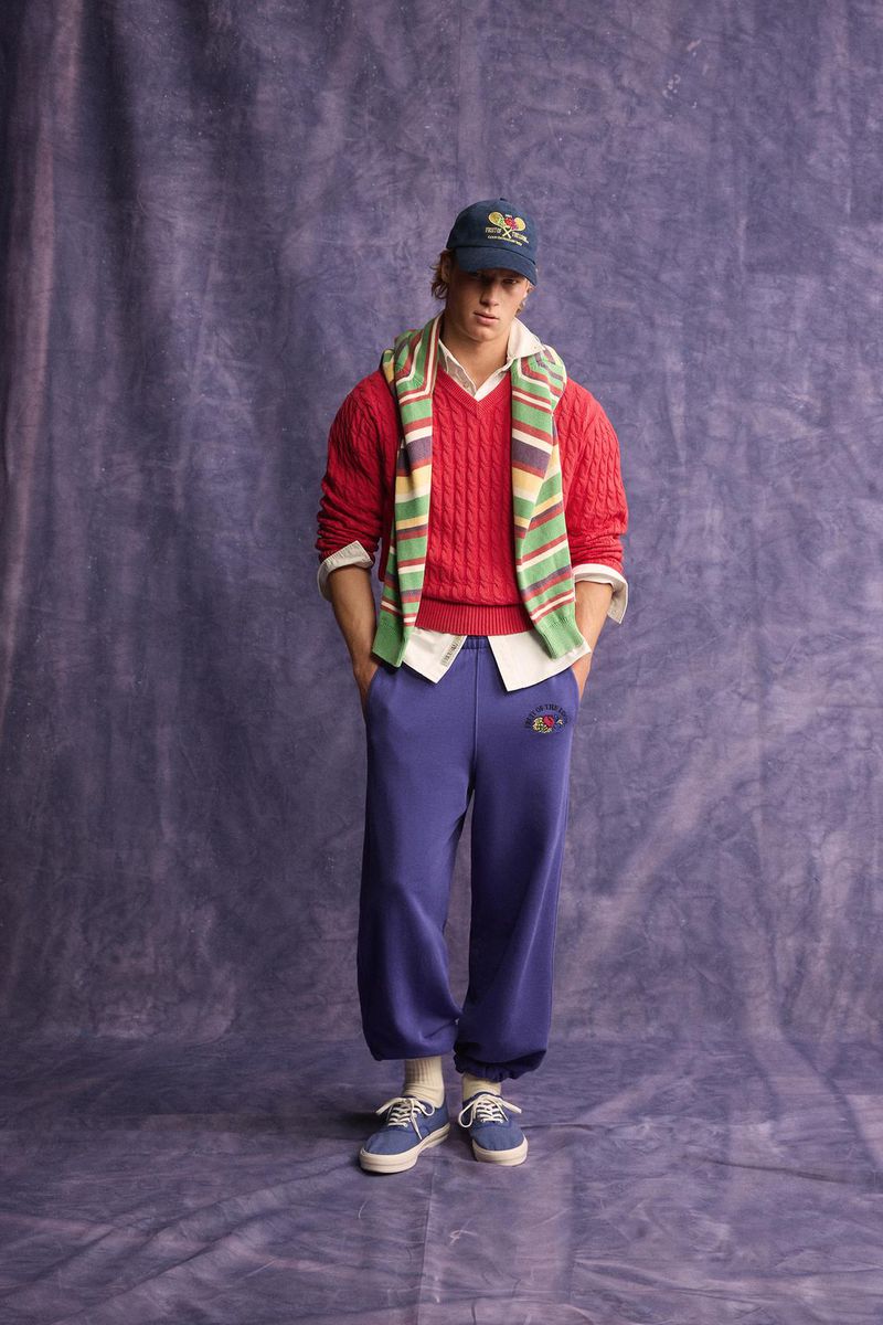 Model wearing a red cable knit sweater, striped scarf, cap, and blue sweatpants standing against a mottled purple backdrop.