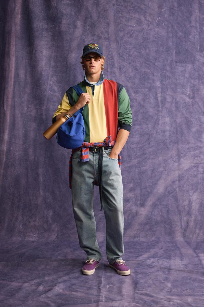 Model wearing a color-blocked rugby shirt and blue jeans standing against a purple textured backdrop for Zara.