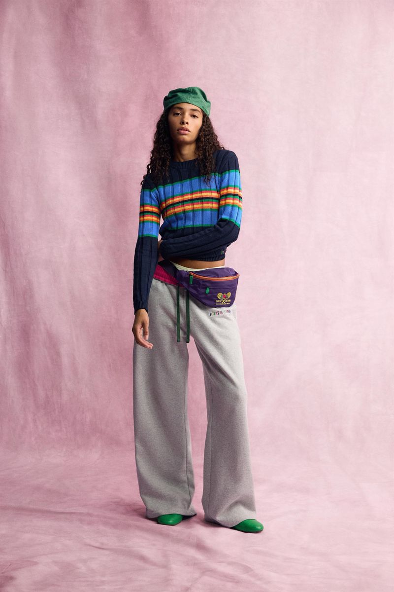 Model wearing striped sweater, grey sweatpants, green head covering, and fanny pack against textured pink backdrop