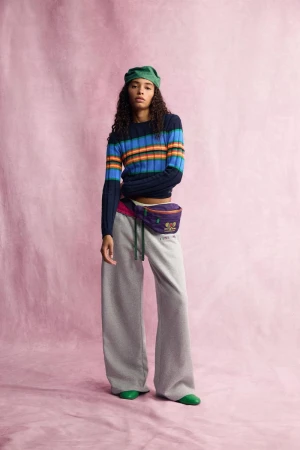 Model wearing striped sweater, grey sweatpants, green head covering, and fanny pack against textured pink backdrop