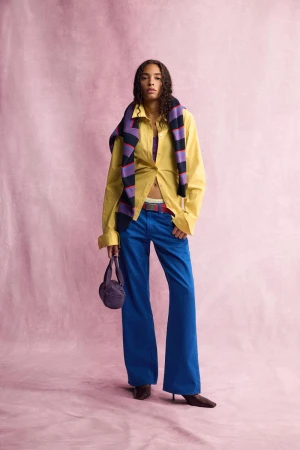 Model standing against a pink textured backdrop wearing a yellow shirt, blue trousers, and a striped sweater draped over shoulders.