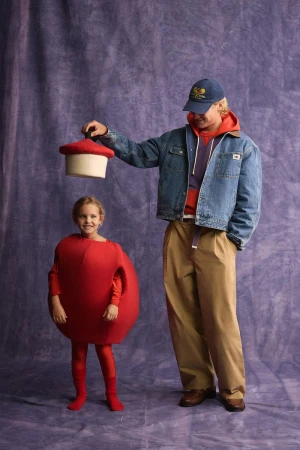 A man in a denim jacket holding a red prop above a smiling child wearing a red fruit costume against a mottled purple backdrop.