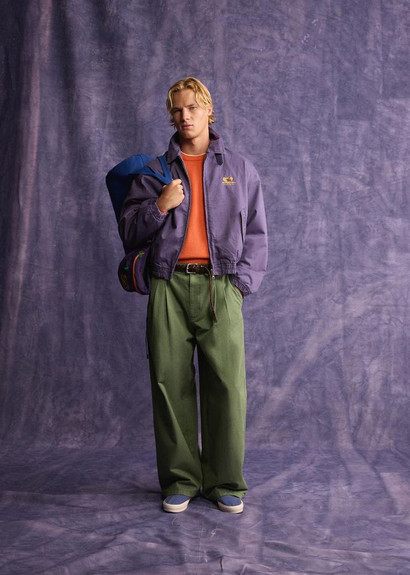 Blonde male model wearing a purple jacket, orange sweater, and green trousers carrying a blue duffel bag against a textured backdrop