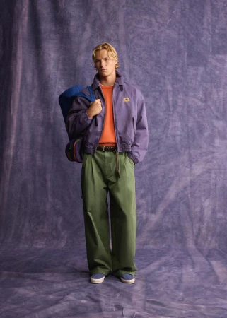 Blonde male model wearing a purple jacket, orange sweater, and green trousers carrying a blue duffel bag against a textured backdrop