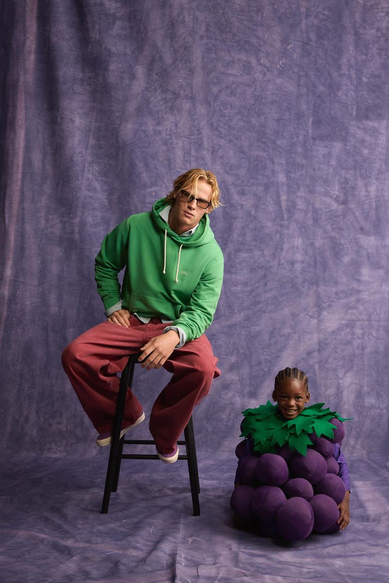 Man in a green hoodie seated on a stool next to a child wearing a purple grape costume against a textured purple backdrop.