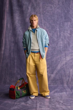 Model standing against a purple textured backdrop wearing layered casual clothing and a duffel bag on the floor
