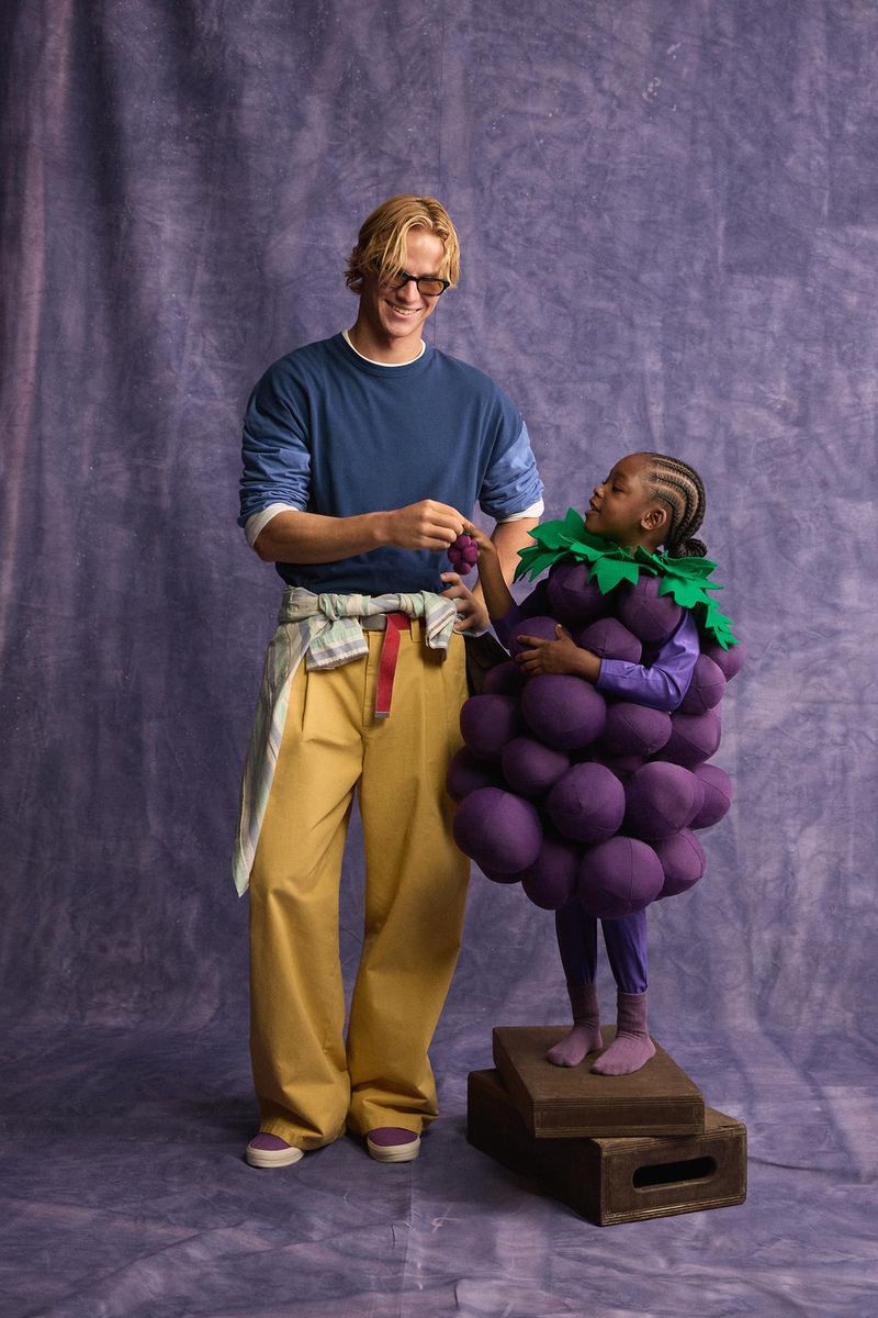 Man in blue shirt and yellow trousers standing next to a child wearing a purple grape costume against a textured backdrop