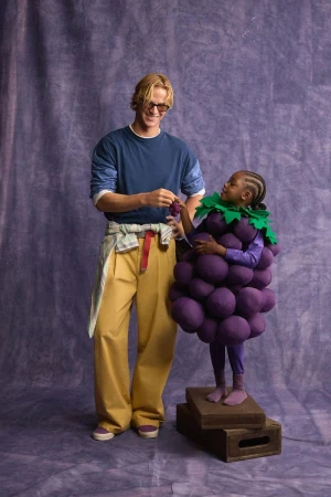 Man in blue shirt and yellow trousers standing next to a child wearing a purple grape costume against a textured backdrop