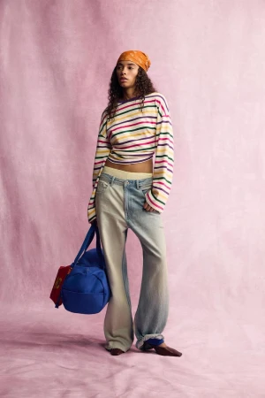 Model wearing a striped long-sleeve top and light-wash jeans holding a blue duffel bag against a pink textured backdrop