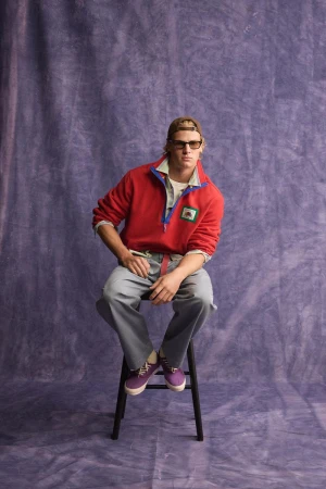 Male model wearing a red pullover, sunglasses, and cap seated on a stool against a textured purple backdrop.