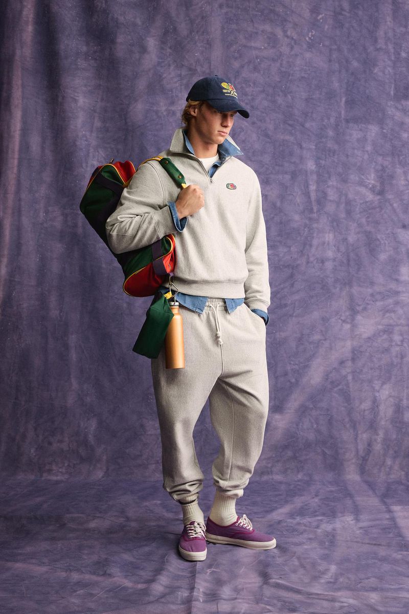 Model wearing a grey sweatshirt and matching sweatpants holding a colourful duffel bag against a purple textured backdrop