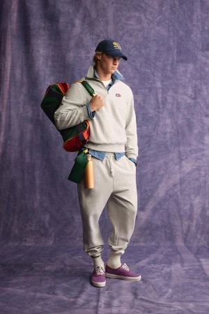 Model wearing a grey sweatshirt and matching sweatpants holding a colourful duffel bag against a purple textured backdrop