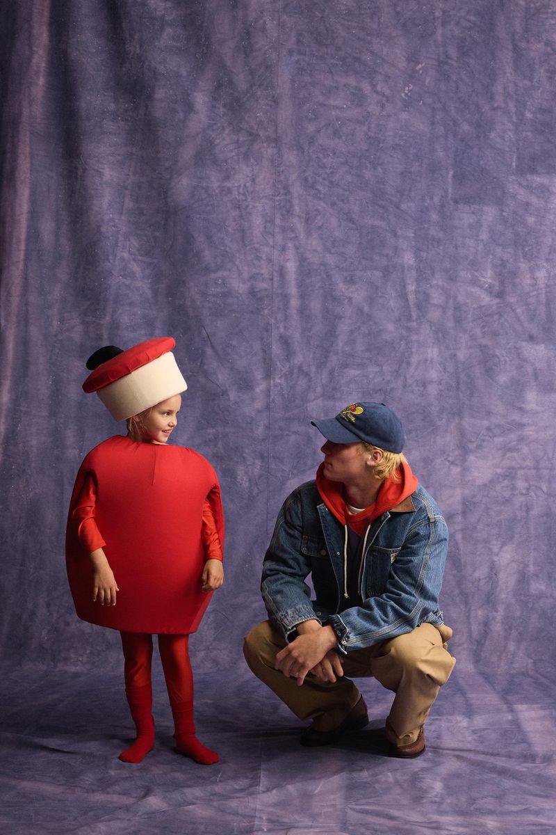 A young girl in a red costume and a man in a denim jacket squatting against a purple textured studio backdrop.