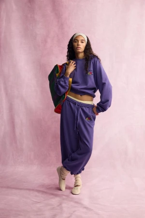 Model wearing purple sweatpants and matching sweatshirt holding a duffel bag against a textured pink backdrop for Zara.