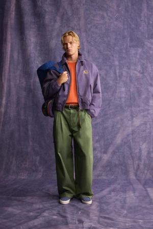 A blonde male model standing against a textured purple backdrop wearing a purple jacket, orange sweater, green trousers, and a blue shoulder bag.