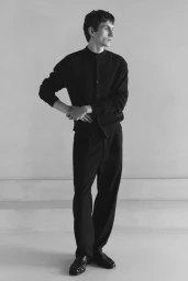 Full-length portrait of a male model wearing a dark buttoned cardigan and trousers against a textured plain wall.