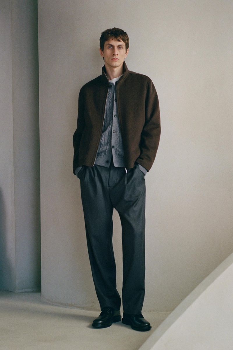 Full-length portrait of a male model wearing a brown zip jacket over a grey cardigan and dark trousers against a plain wall