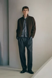 Full-length portrait of a male model wearing a brown zip jacket over a grey cardigan and dark trousers against a plain wall