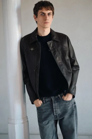 Model in dark leather jacket and grey jeans leaning against a white column in an interior setting