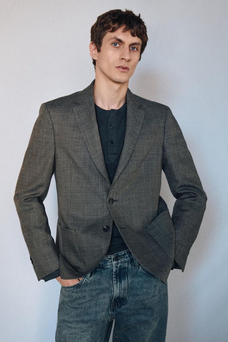 A man wearing a grey plaid blazer over a dark henley shirt and blue jeans against a neutral wall.