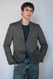 A man wearing a grey plaid blazer over a dark henley shirt and blue jeans against a neutral wall.