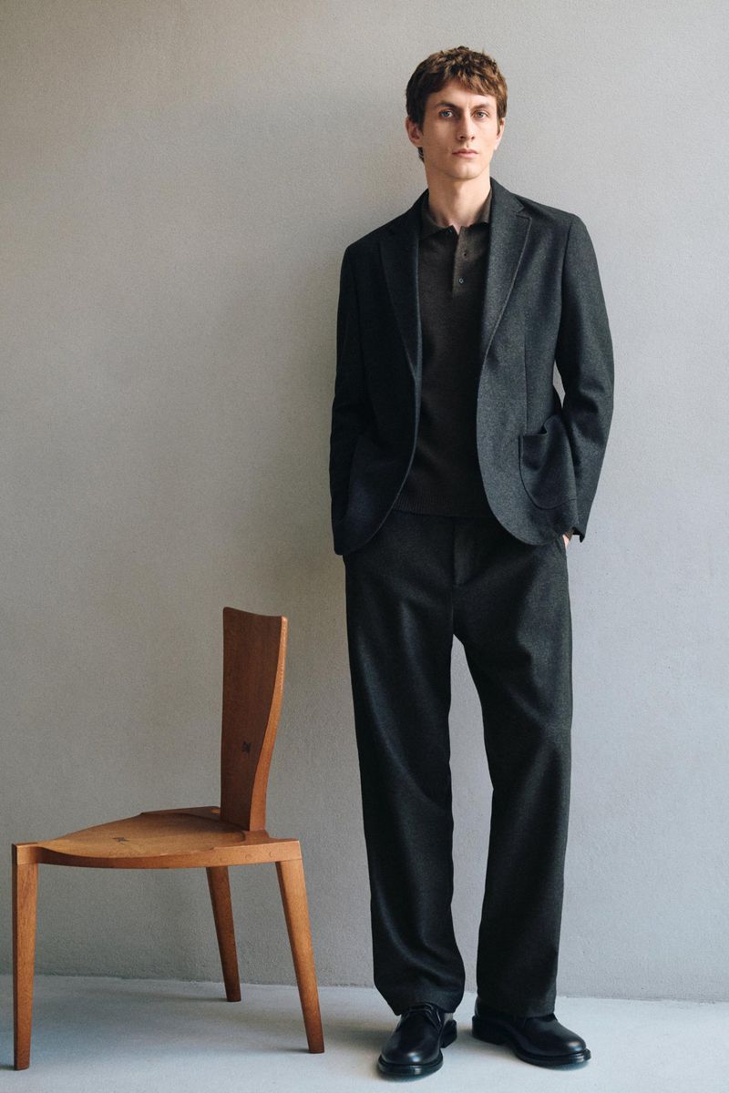 Full-length portrait of a male model wearing a dark suit and polo shirt standing beside a wooden chair.