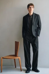 Full-length portrait of a male model wearing a dark suit and polo shirt standing beside a wooden chair.
