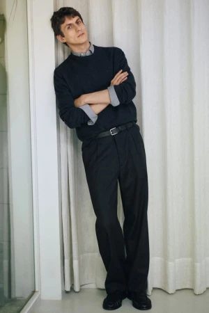 A man leaning against a white wall with folded arms wearing a dark crewneck sweater over a patterned collared shirt and dark trousers.